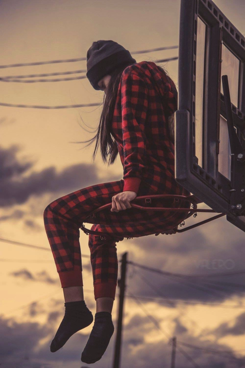 A person in red plaid and a dark beanie sits in a suspended window frame against a sunset sky.