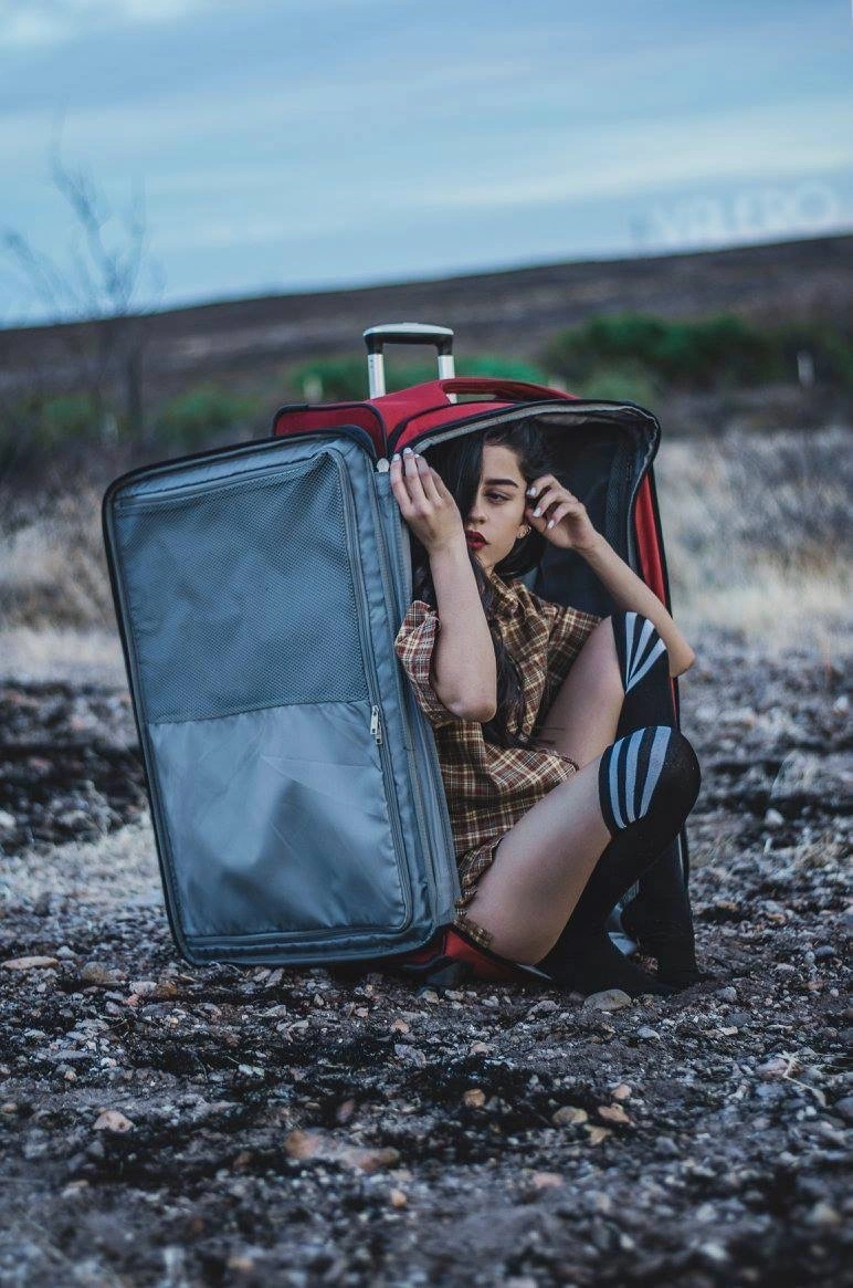 A woman curls up inside an open red suitcase on gravel outdoors.