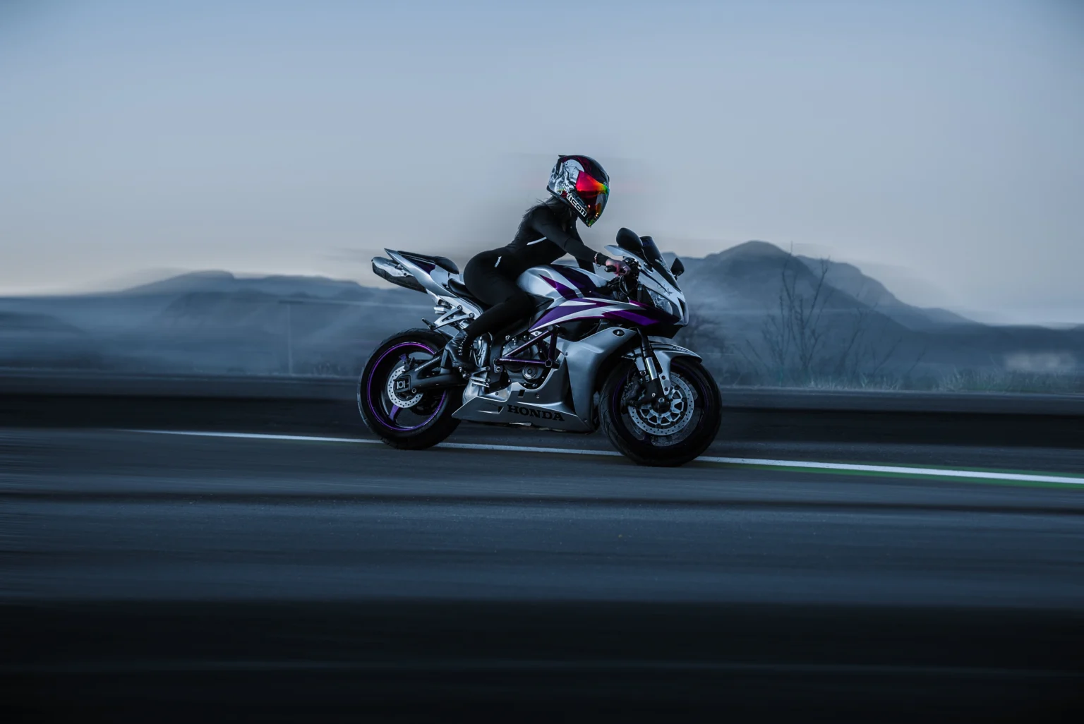 A helmeted motorcyclist rides a purple and white sportbike along a road with mountains behind.