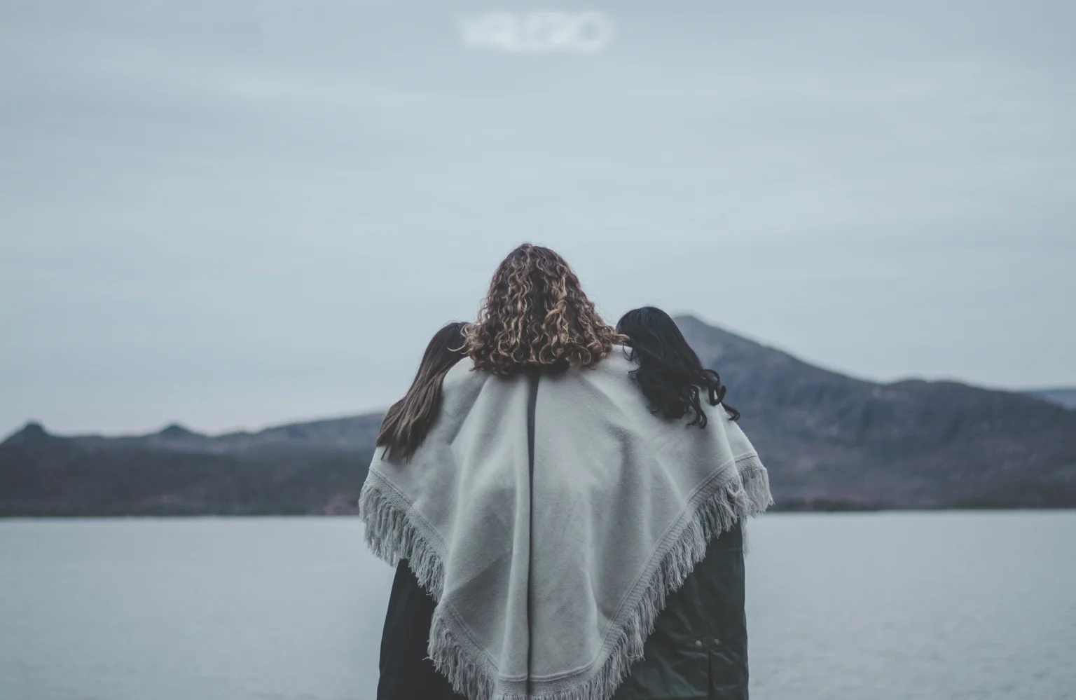 People wrapped together in a pale blanket beside a lake, facing distant mountains.
